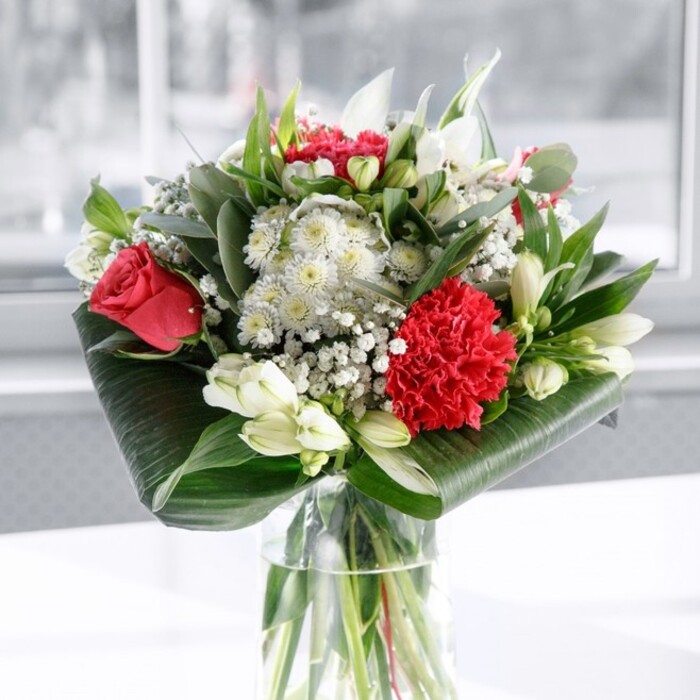 A beautifully composed bouquet in rich reds and soft whites stands in a clear cylindrical vase, the kind you might find gracing a dining table in a Upton Park terrace house after a same-day delivery. Through the transparent glass, you can see the neatly cut green stems fanning out in clear water, a quiet detail that suggests care, freshness, and a professional touch. Above the rim, the flowers form a generous dome: deep red roses with velvety, unfolding petals rise gracefully, while a large, frilled crimson carnation towards the front adds a sculptural burst of texture. Threaded between these bold red notes are white blooms in varying forms-slender, speckled Alstroemeria, creamy white lily buds still closed and brimming with promise, and soft puffs of gypsophila that appear like tiny pearls, lending a delicate, almost bridal air. The arrangement is framed by lush greenery. At the base, broad, dark green tropical leaves are artfully curled and wrapped, creating a smooth, structured collar that holds the bouquet together. Above, eucalyptus-like foliage and pointed leaves in differing shades of green weave in and out, adding movement and depth without overwhelming the blossoms. The scene is set on a spotless white surface with a bright, gently blurred background that suggests daylight filtering through a window, perhaps overlooking nearby West Ham Park. The lighting is soft and even, allowing every petal, leaf, and stem to be clearly defined. As a whole, the bouquet feels modern yet timeless-perfect as a centrepiece for family celebrations, anniversaries, or quiet moments of gratitude shared in homes and workplaces across Upton Park.