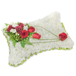 A solemn and meticulously crafted memorial pillow, this rectangular cushion of blooms reads as a quiet homage. The entire surface is densely carpeted with small, pristine white chrysanthemum heads, packed closely so the pillow looks as soft and consoling as a blanket of fresh snow; the petals create a fine, velvety texture that suggests rest and purity. Around the gently rounded edge a slim band of pale green ribbon and occasional sprigs of foliage peek out, giving a neat, finished outline that whispers of careful handwork rather than ostentation.
At the upper left a cluster of deep, blood-red roses and rich burgundy carnations gathers like a heart of remembrance. Their velvety petals and ruffled layers form a dramatic, dignified contrast against the white field. Interspersed among those blooms are small white accent flowers-freesias or tiny spray chrysanthemums-that catch the light and add delicate points of brightness. Behind and between the flowers, feathery fern fronds and glossy green leaves give weight and depth, while thin, arching strands of bright green bear-grass sweep diagonally across the cushion, creating a gentle line of movement from the main spray toward the lower right.
The sweep culminates in a single, perfectly formed red rose resting on the pillow's lower corner, as if cradled by those curving stems-a simple symbol of enduring love. In soft daylight the arrangement reads both refined and reassuring; its scent is understated, a clean floral hush. This traditional sympathy cushion, often chosen for funeral services and memorial displays in Upton Park chapels and local wakes, conveys comfort, respect and steady affection in moments of farewell.